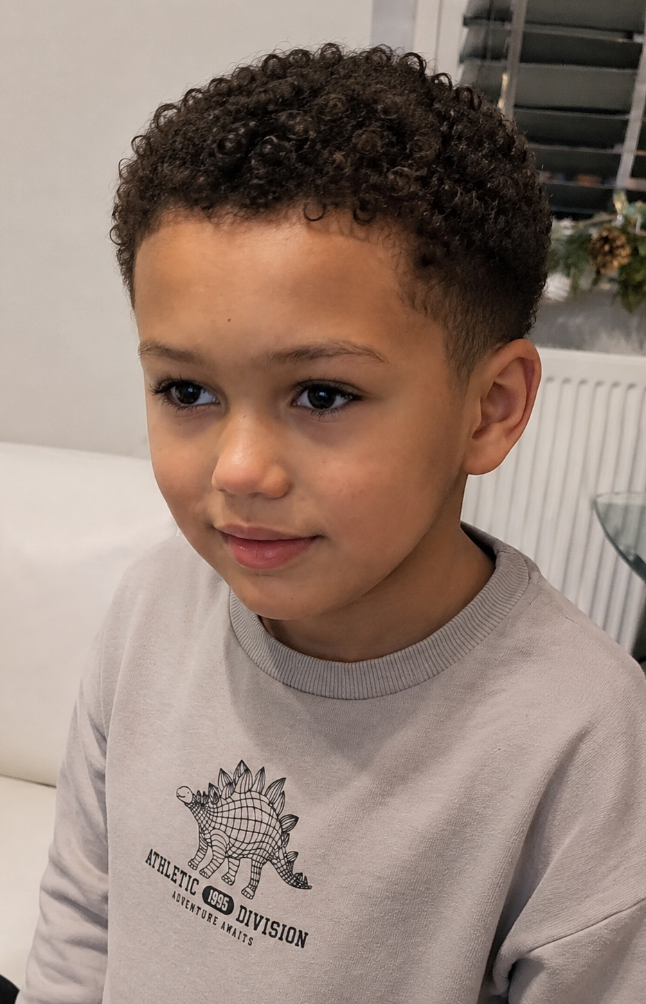 A young boy with curly hair, wearing a grey sweater, looking thoughtfully off to the side.